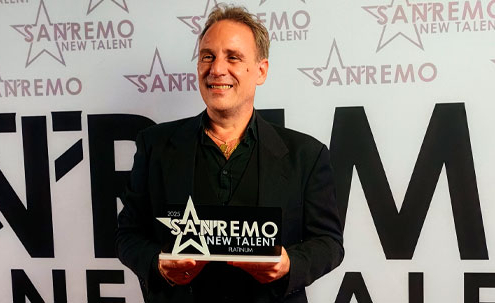 Roberto Quintavalle sorride con orgoglio, tenendo in mano la targa di vincitore della categoria Platinum sul palco di Sanremo New Talent 2025, davanti al backdrop ufficiale del concorso.