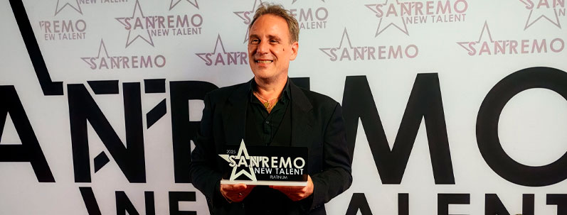 Roberto Quintavalle sorride con orgoglio, tenendo in mano la targa di vincitore della categoria Platinum sul palco di Sanremo New Talent 2025, davanti al backdrop ufficiale del concorso.
