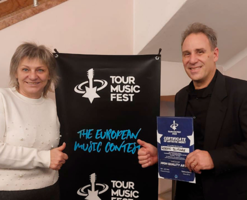 Roberto Quintavalle e Liuba posano ai lati del banner del Tour Music Fest 2025 subito dopo la prima performance; Roberto elegante in abito di scena con attestato di merito "High Quality Artist".