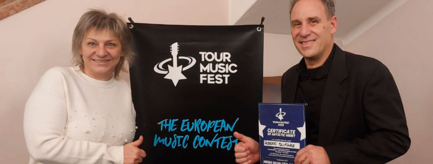 Roberto Quintavalle e Liuba posano ai lati del banner del Tour Music Fest 2025 subito dopo la prima performance; Roberto elegante in abito di scena con attestato di merito "High Quality Artist".